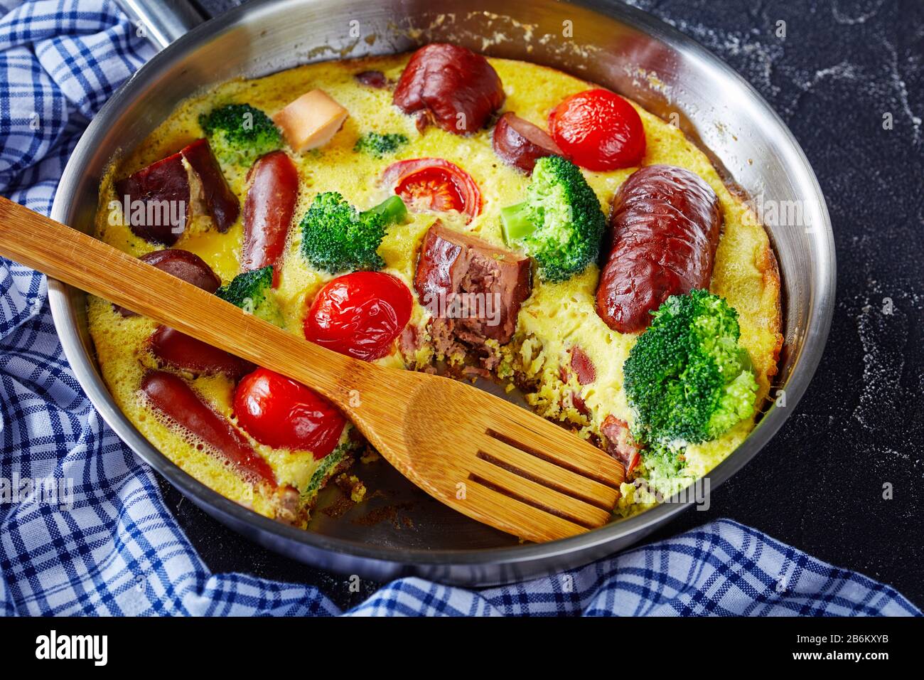 Frittata mit Würstchen, Brokkoli, Kirschtomaten, geschmolzenem Käse in einem Skillet mit Spachtel auf einem Betontisch, horizontale Ansicht von oben, Nahaufnahme Stockfoto