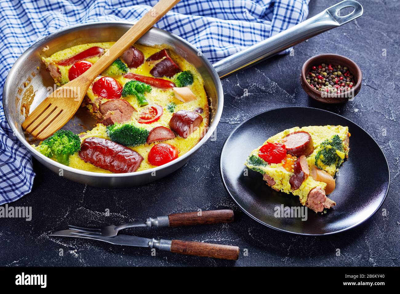 Nahaufnahme von Omelette mit Würstchen, Brokkoli, Kirschtomaten, geschmolzenem Käse in einem Skillet auf einem Betontisch und einem Teil auf einem Teller, horizontale Ansicht Stockfoto