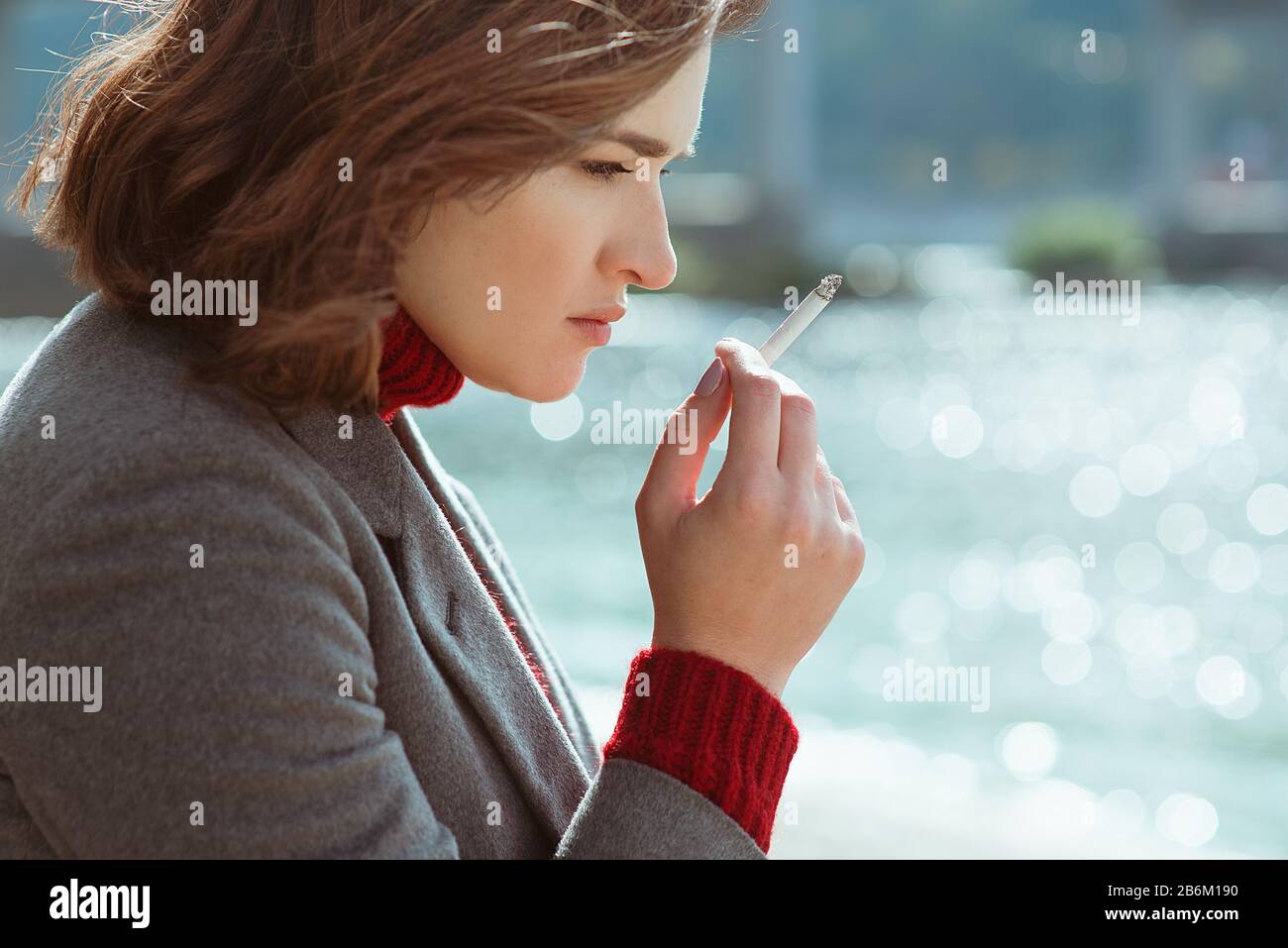 Schöne stilvolle traurig stressig junge Frau in Pullover und Mantel Rauchen Zigarette in der Nähe des Flusses Stockfoto