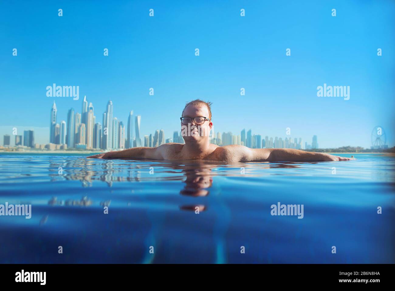 Porträt eines fröhlichen entspannten europäischen kaukasischen Rotkopfmannes in Gläsern im Swimmingpool auf dem modernen Dubai (Vereinigte Arabische Emirate) und Meereshintergrund Stockfoto