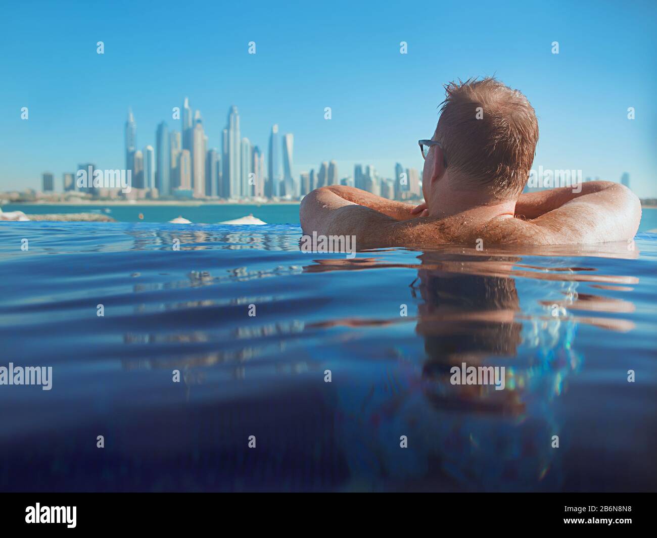 Porträt eines entspannten europäischen kaukasischen Rotkopfmannes mit Haaren zurück in Gläsern im Schwimmbad auf dem modernen Dubai (Vereinigte Arabische Emirate) und Meeresbac Stockfoto