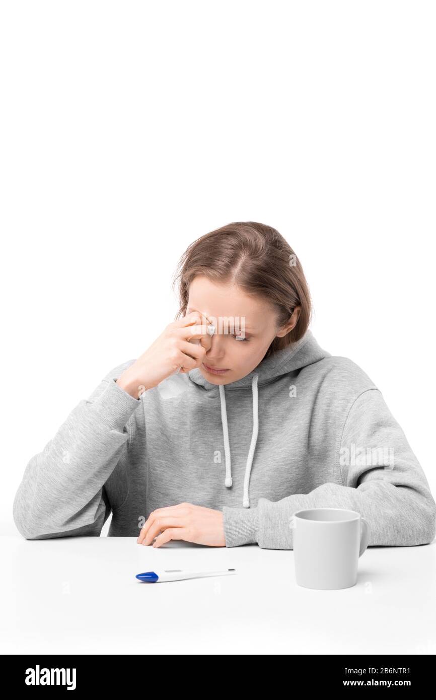 Kranke junge Frau, die mit Digitalthermometer und Becher am Tisch sitzt und Tränen wischt, während sie wegen Krankheit weint Stockfoto