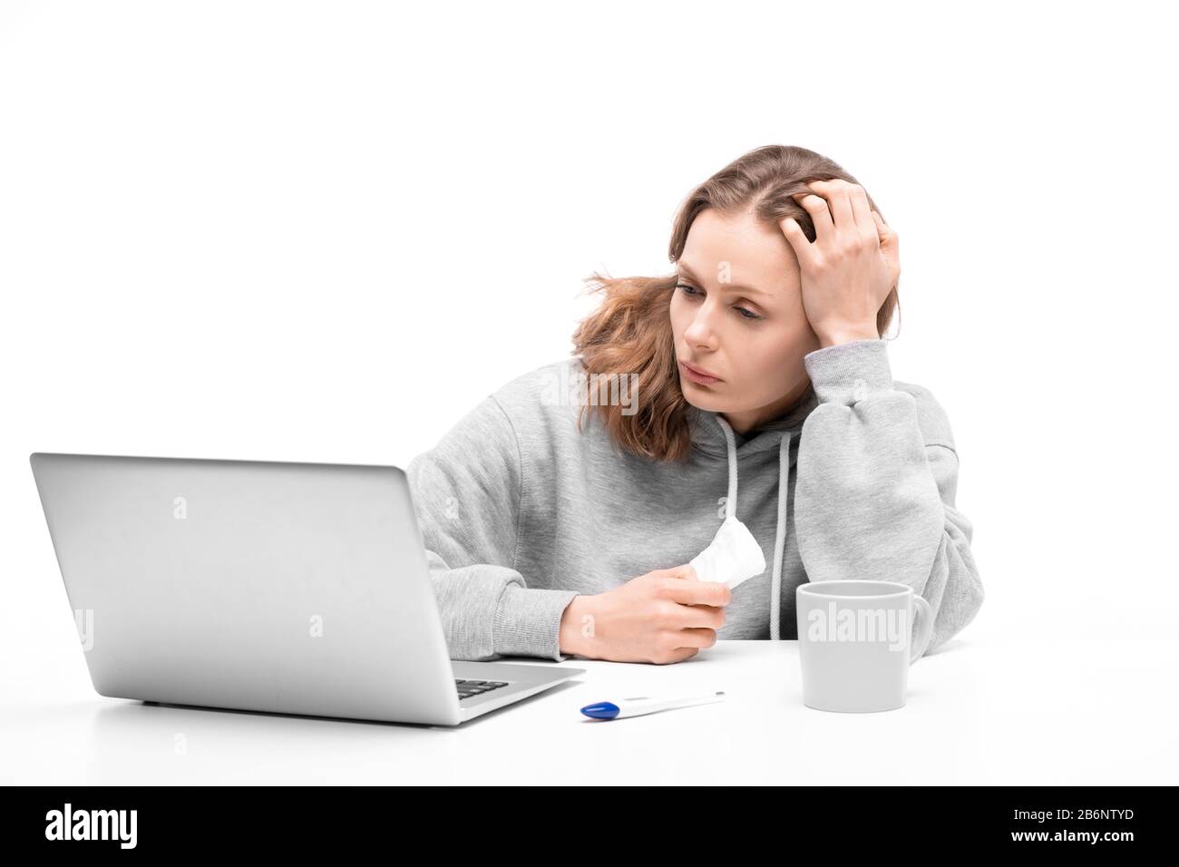 Junge Frau in Hoodie sitzt am Tisch und hält Serviette, während sie über die Symptome im Internet mit Laptop liest Stockfoto