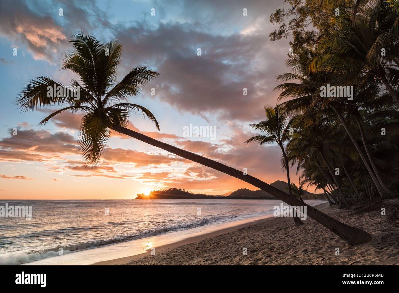 Eine kultige Kokospalme hängt schräg über einer typischen Sonnenaufgang-, tropischen Strandszene mit Insel im Hintergrund und Kokospalmenlinie. Stockfoto