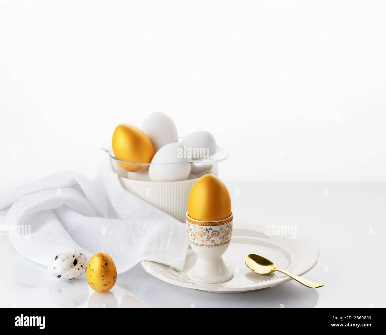 Frühstücks-Konzept für Ostern. Weiches gekochtes Ei auf einem weißen Teller mit goldenen und weißen Ostereiern auf weißem Grund. Weicher Fokus. Stockfoto