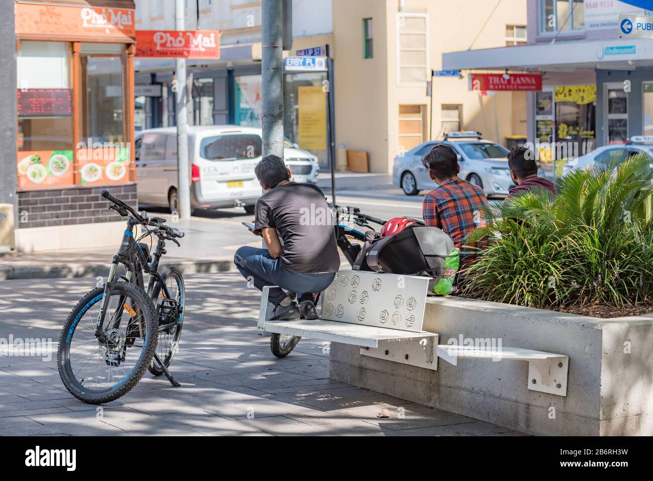 Fahrradkuriere sitzen und warten auf Arbeit in der Nähe lokaler Restaurants in einem kleinen Park in Parramatta, New South Wales, Australien Stockfoto