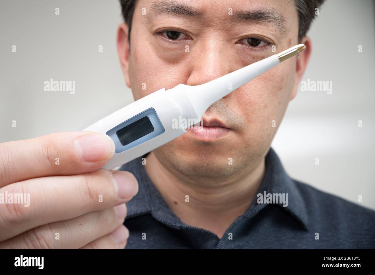 Ein asiatischer Mann mittleren Alters, der das Thermometer ängstlich in der Hand betrachtet. Stockfoto