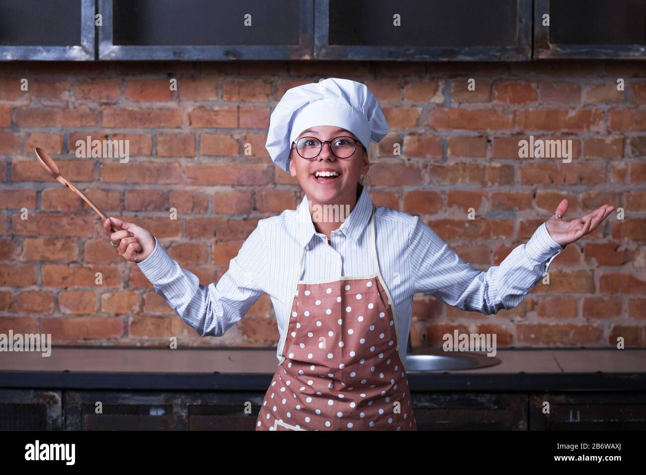 Lächelndes Mädchen im Teenager-Anzug mit einer Schürzenkappe, hält einen Löffel und zuckt. Aus der Küche im Hintergrund. Stockfoto