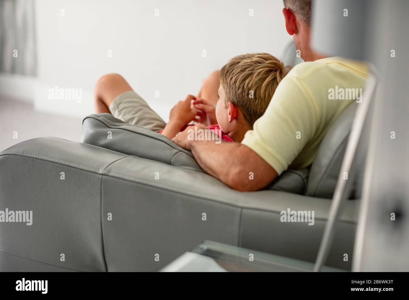 Eine Rückansicht eines jungen Jungen, der mit seinem Vater auf einer Couch aufliegt und Fernsehen sieht, trägt sie legere Kleidung. Stockfoto
