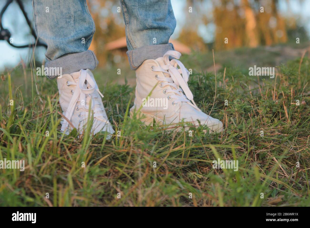 Nahaufnahme der Füße von Teenagern in modernen und trendigen weißen Sneakern und gerollten Jeans. Sonniger Sommertag, grüner Gras Hintergrund. Stockfoto