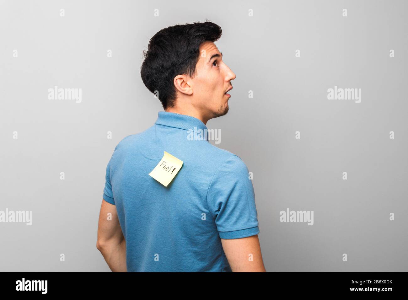 Dumm aussehender Mann in blauem Hemd mit gelber Haftnotiz mit Wortnarren auf dem Rücken stumm nach oben blickend Stockfoto