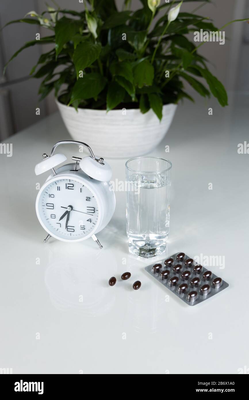 Weißer Wecker und medizinische Pillen mit einem Glas Wasser auf dem Tisch. Gesundheitswesen und Medizin. Vitamine und Mineralstoffe in Kapseln. Stockfoto