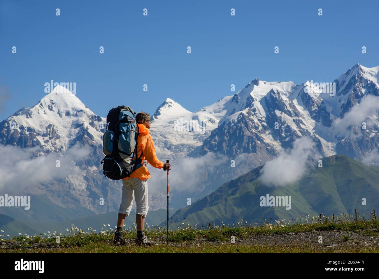 Frau (Backpacker) mit Sonnenbrille und Stöcken betrachtet schneebedeckte Berge. Touristen liegt auf der Alpenwiese. Stockfoto