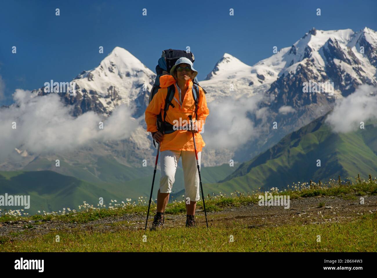 Frau (Backpacker) mit Sonnenbrille und Stöcken gegen Berge mit Schneehintergrund. Touristen liegt auf der Alpenwiese. Tviber Mount ist i Stockfoto