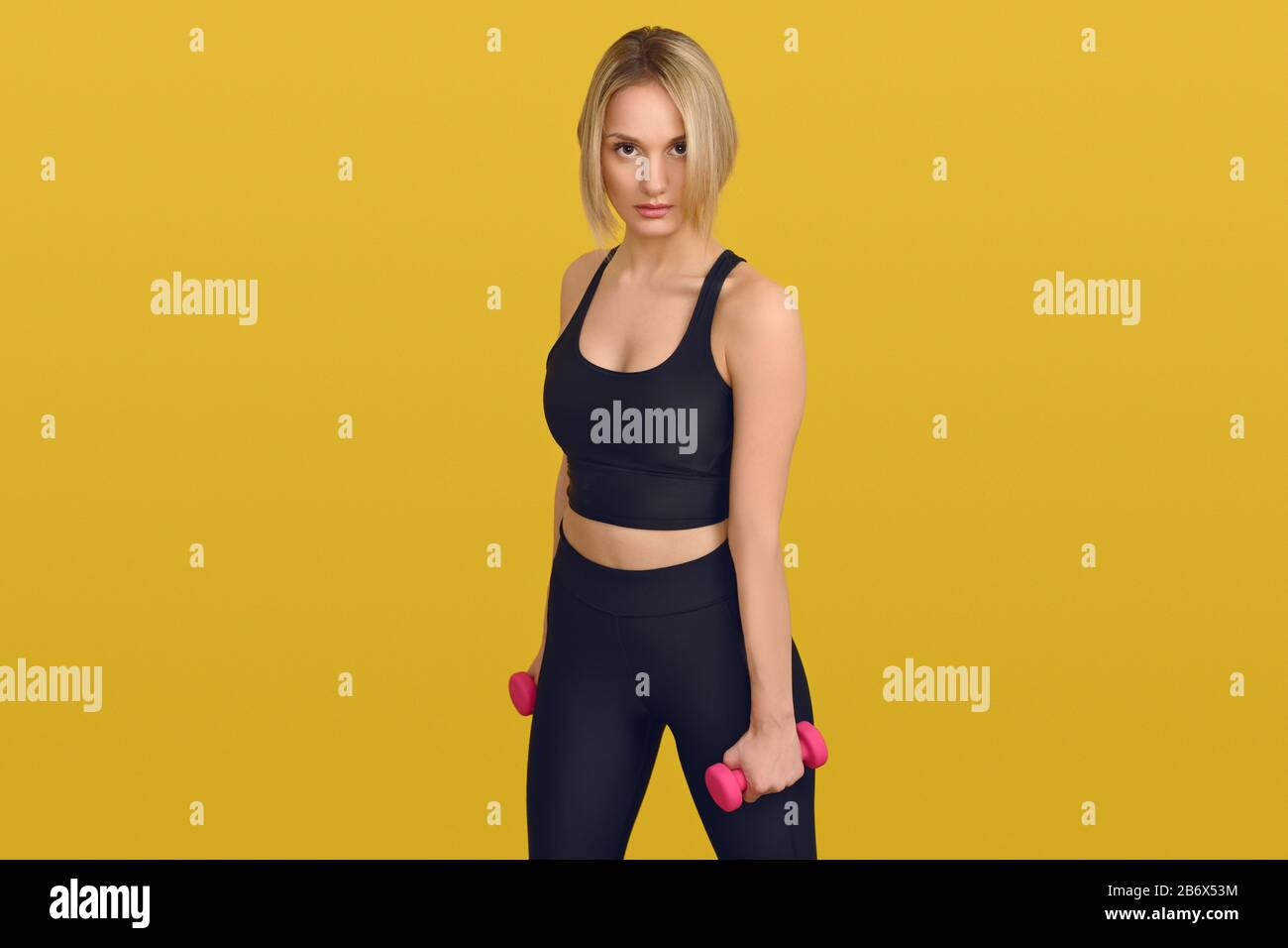 Hübsche blonde Frau in schwarzem Trainings-Shirt mit kleinen pinkfarbenen Hanteln in den Händen, mit Blick auf die Kamera. Nahaufnahme vorne Hochformat gegen einfarbige gelbe Rückseite Stockfoto