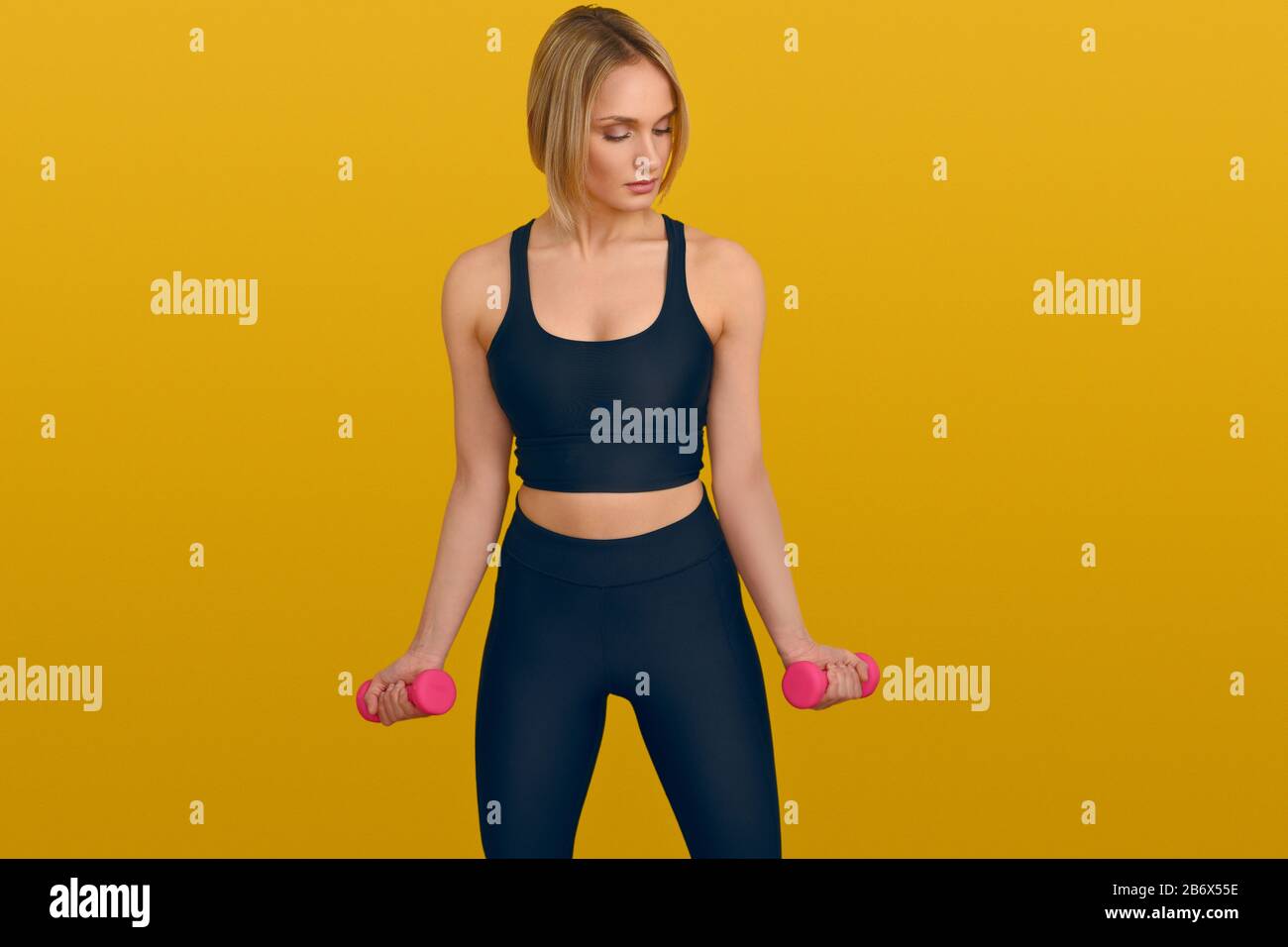 Junge und schöne blonde Frau in schwarzer Trainingskleidung mit pinkfarbenen Hanteln in den Händen vor einfarbig gelbem Hintergrund mit Kopierraum, a Stockfoto