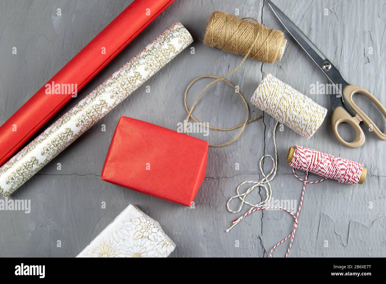 Vorbereitung auf die Verpackung von Geschenken, auf dem Tisch aufgelegtes rot-weißes Verpackungspapier, Fäden verschiedener Farben für Wicklung und Schere Stockfoto
