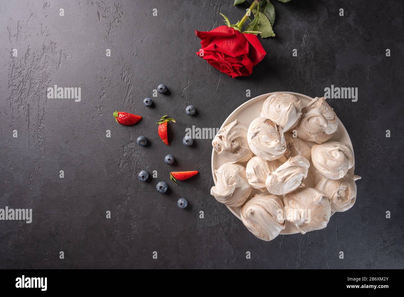 Hausgemachtes, beliebtes Dessertmergue auf einem weißen Teller auf schwarzem Hintergrund. In der Nähe liegen Erdbeeren und Blaubeeren und rote Rosenblüblume Stockfoto