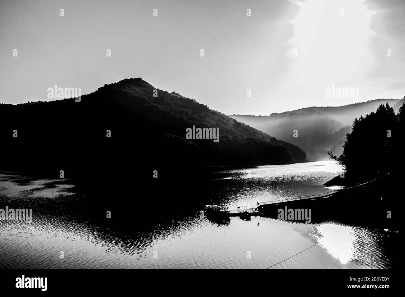 Schwarz-weiße Sonnenzusammensetzung steigt über einen nebligen See mit Bergwald im Hintergrund und Boot an der Pier im Vordergrund Stockfoto