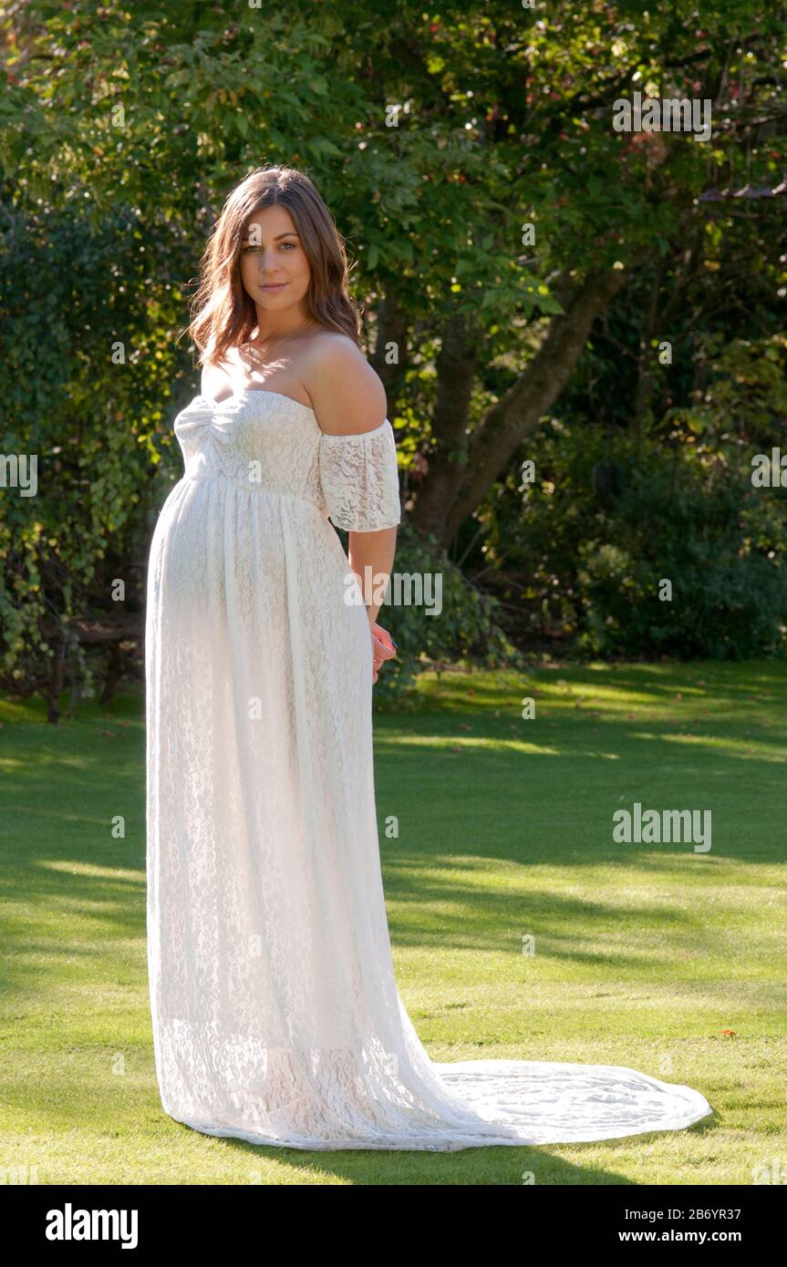 Schöne schwangere junge Frau, die ein langes weißes Kleid trägt und ihr Baby streichelt Stockfoto