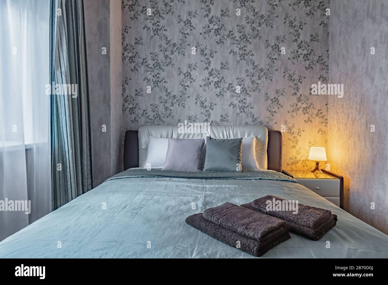 Frisches, sauberes, gemütliches Bett in einem kleinen, gemütlichen Schlafzimmer mit warmer Lampe am Abend Stockfoto