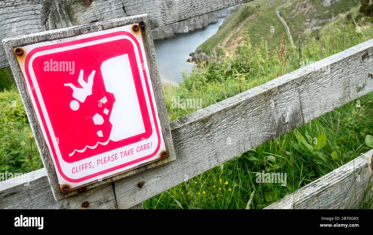 Warnschild an den Smoo Cave Cliffs, Schottland. Ein bebildertes Zeichen, das vor einer schieren Felswand warnt. Der Tropfen ist durch den Zaun sichtbar. Stockfoto
