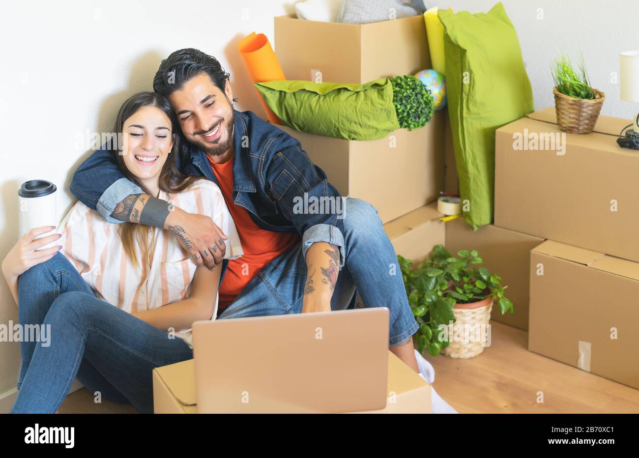 Glückliches junges Paar, das zum ersten Mal nach Hause zieht - Mann und Frau haben Spaß mit dem Computer in der nächsten Kartonbox im neuen Haus Stockfoto