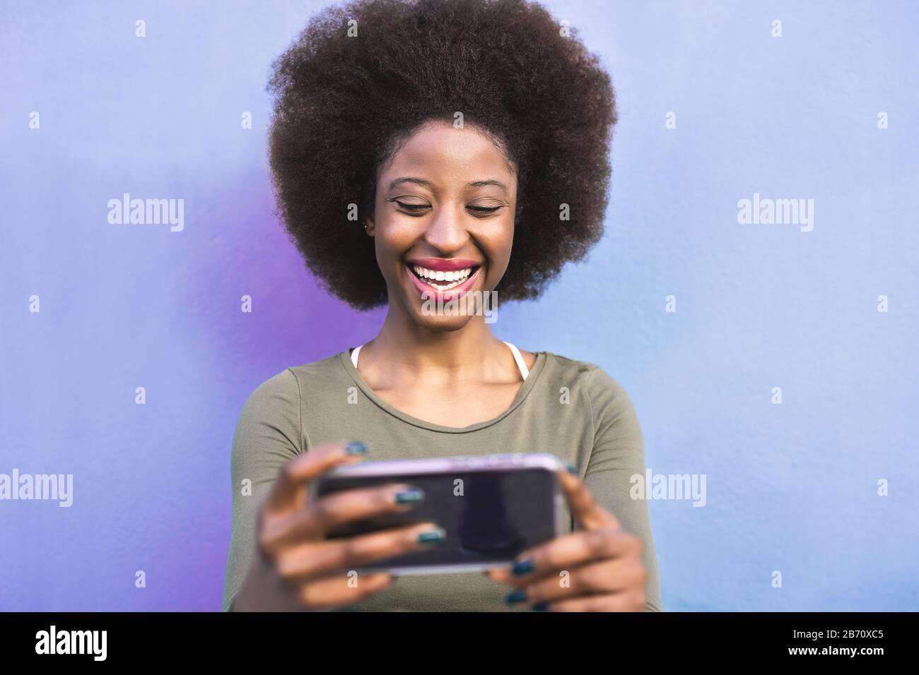 Fröhliches Afro Mädchen mit Handy im Freien - Lächelnde, soziale Einflussnehmerin, die mit neuen Trends Technologie Apps Spaß hat Stockfoto