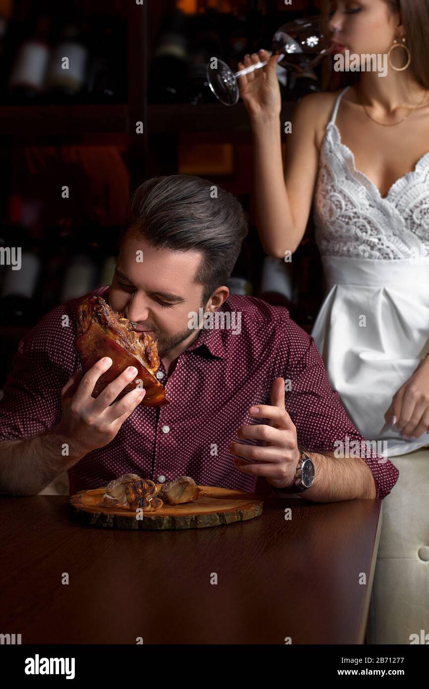Der Kerl isst eifrig einen Schweineknäuel. Dame, die auf einer Couch sitzt, mit einem Glas Rotwein, auf dunklem Hintergrund in einem Restaurant. Stockfoto