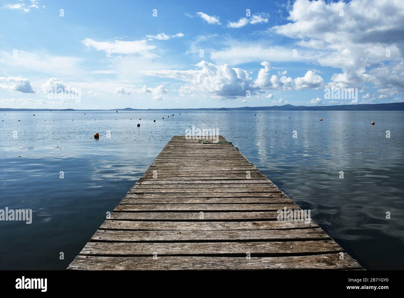 Hölzerne Anlegesteg für Boote vor blauem Himmel mit einigen Wolken. Bojen. Hügeln im Hintergrund. Bolsena See, Italien. Stockfoto