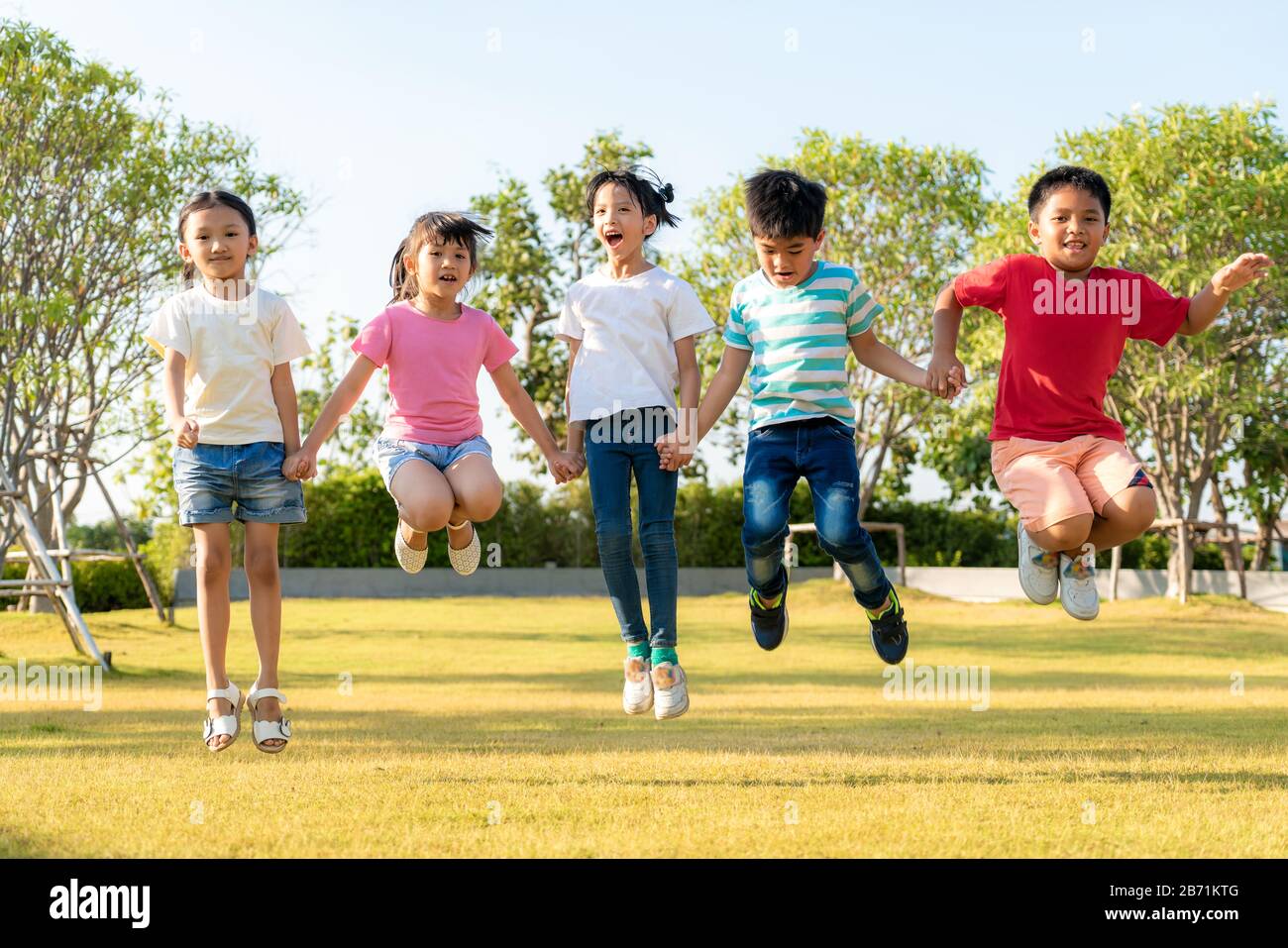Große Gruppe von fröhlichen asiatischen Kindergartenfreunden, die an einem sonnigen Tag in legeren Kleidungsstücken in der Stadt p die Hände beim Spielen und Springen zusammenhalten Stockfoto