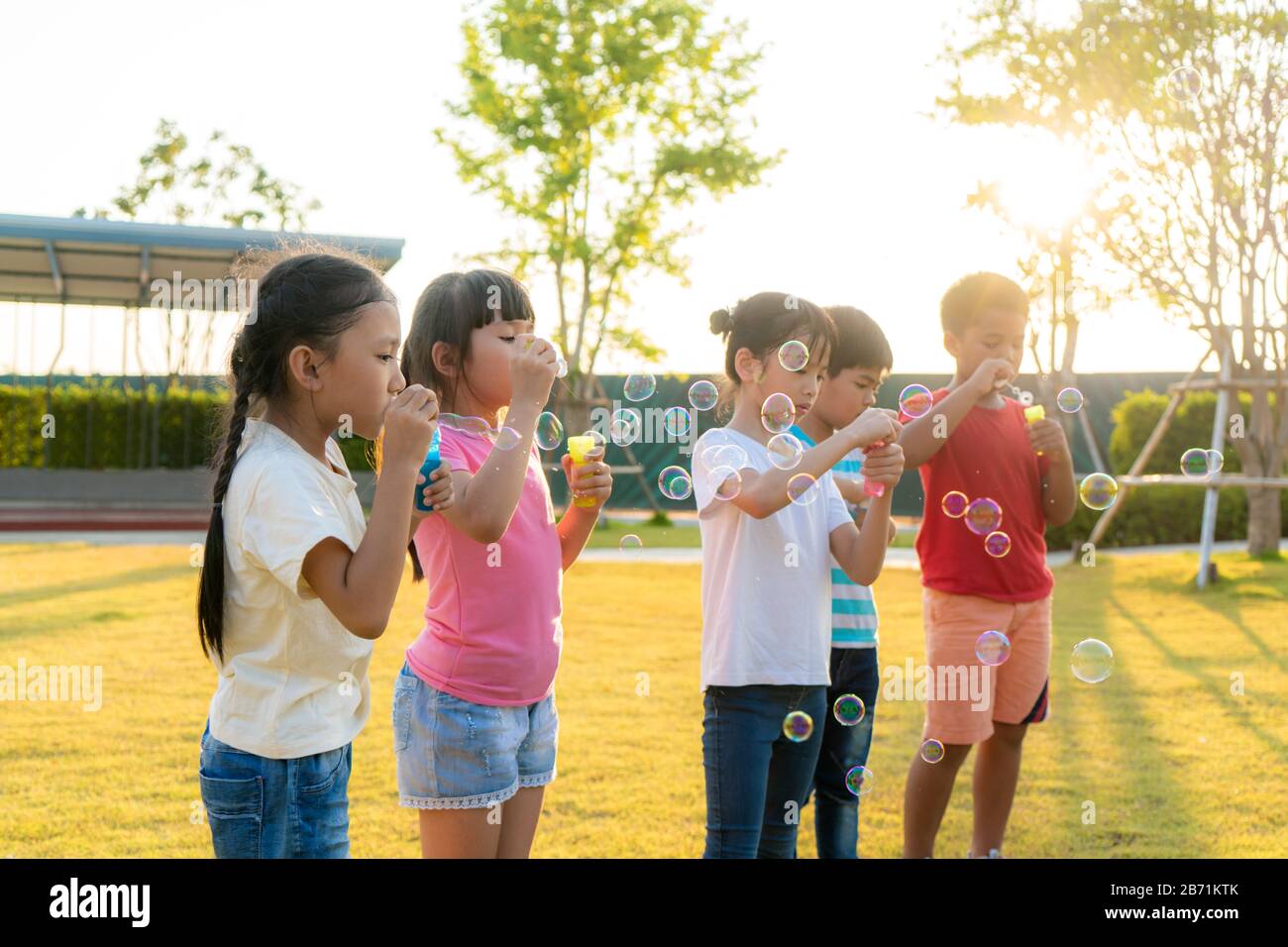 Große Gruppe von fröhlichen asiatischen Kindergartenfreunden, die Blasen im Park auf dem grünen Gras am sonnigen Sommertag zusammen spielen. Stockfoto