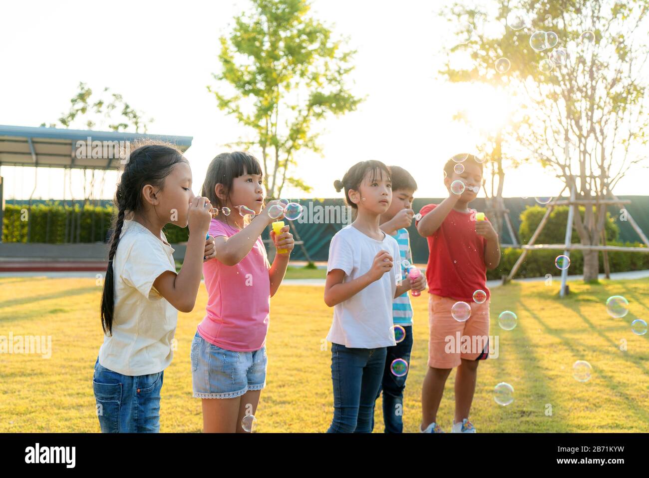 Große Gruppe von fröhlichen asiatischen Kindergartenfreunden, die Blasen im Park auf dem grünen Gras am sonnigen Sommertag zusammen spielen. Stockfoto