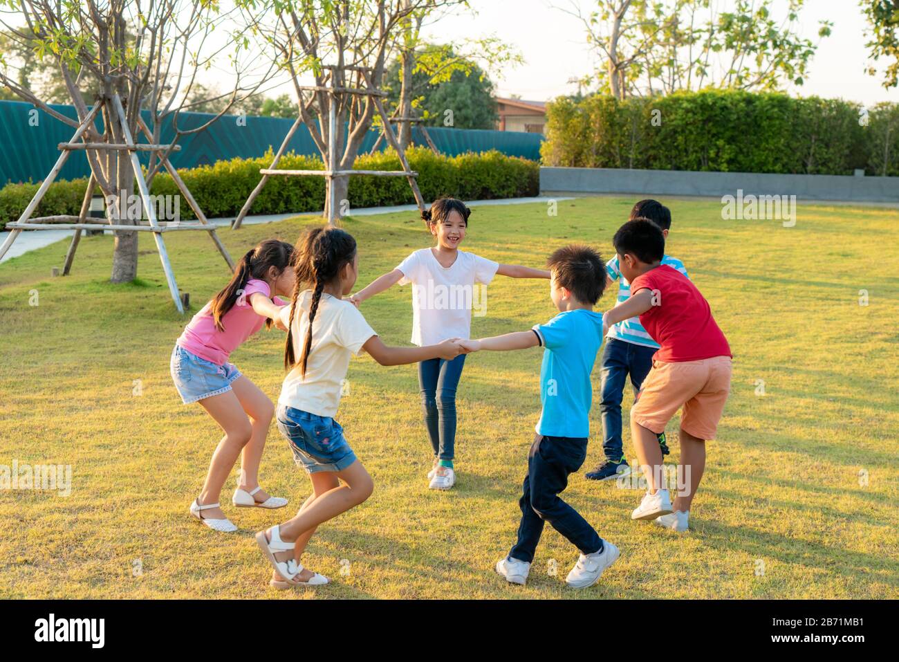 Große Gruppe von fröhlichen asiatischen Kindergartenkindern, die Hände beim Spielen und Tanzen halten, spielen sich rundherum und stehen im Kreis im Park am g Stockfoto