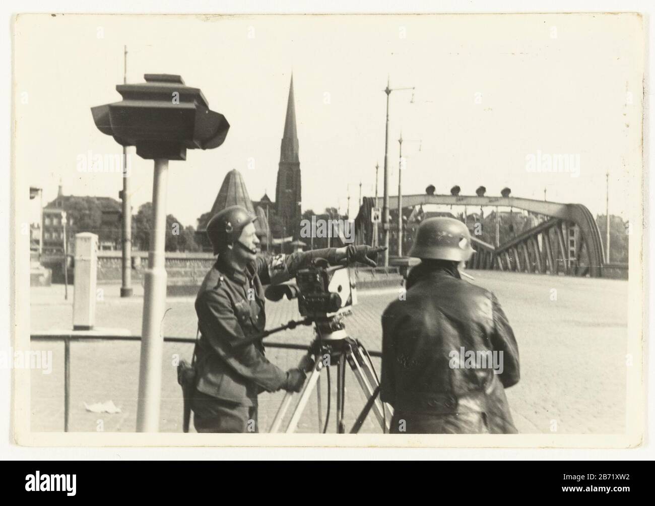 Leden van een Duitse Propaganda Kompagnie Zwei deutsche Mitglieder Der Propaganda Kompagnie drehen Filme auf einer Brücke in einer Stadt. Die Kamera zeigt nach rechts. Beide tragen einen Helm. Sie haben einen Alarm ausgelöst. Neben der Brücke ein Kerktoren. Hersteller : Fotograf: Anonymer Ort Herstellung: Rotterdam Datum: 15. Mai 1940 - ca. 20. Mai 1940 Physische Merkmale: Gelatine silbernes Druckmaterial: Papiertechnik: Gelatine silberfarbenes Druckmaß: H 6,5 cm. B × 10 cm. Betrifft: Filmaufnahmen, Kameramann bei Work Second World, Wenn: 1940 - 1945 Stockfoto