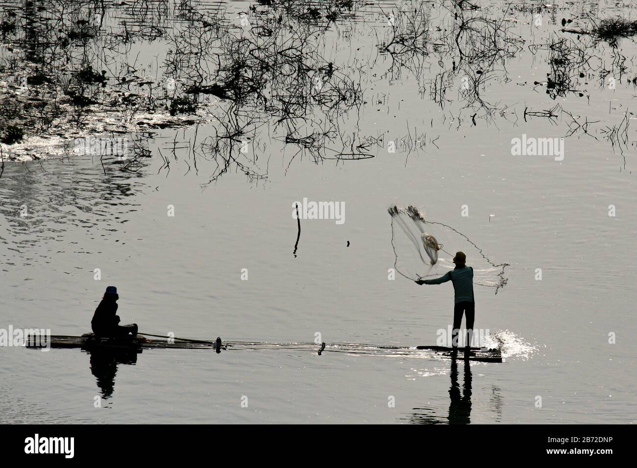 Eine Silhouette von zwei Männern in der Abendsonne auf einem Bambusboot, das Fische durch werfen eines Netzes fängt Stockfoto