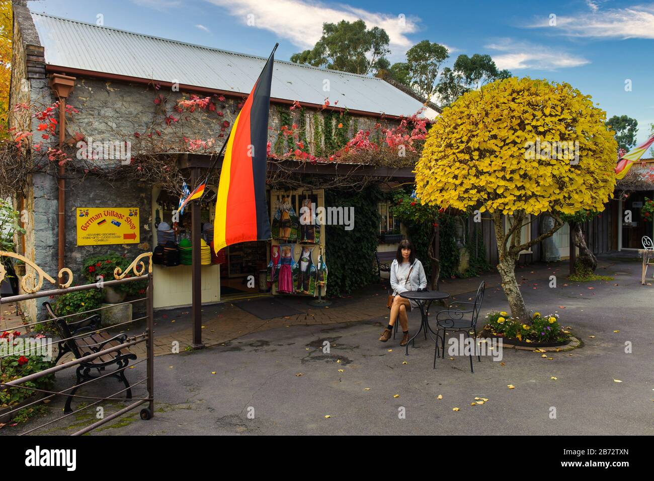 Ein weiblicher Tourist sitzt vor einem alten Steinladen mit deutscher Flagge und wartet auf ihren Kaffee, der von der Herbstflora umgeben ist Stockfoto
