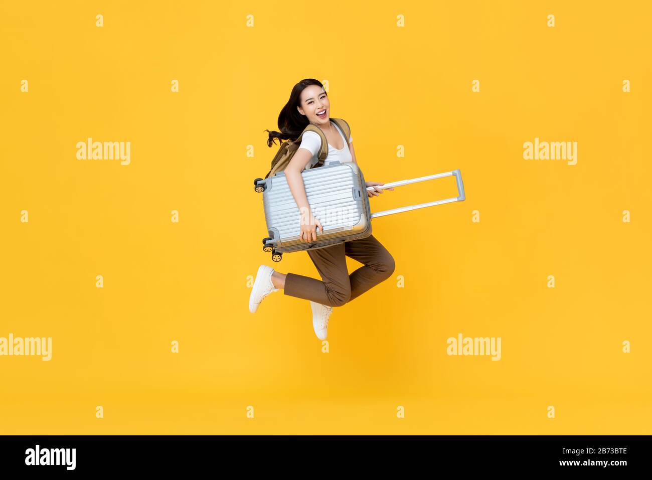 Aufgeregter, schöner Asian Tourist mit Gepäckspringen im Mittelluftstudio, der isoliert auf gelbem Hintergrund gedreht wurde Stockfoto