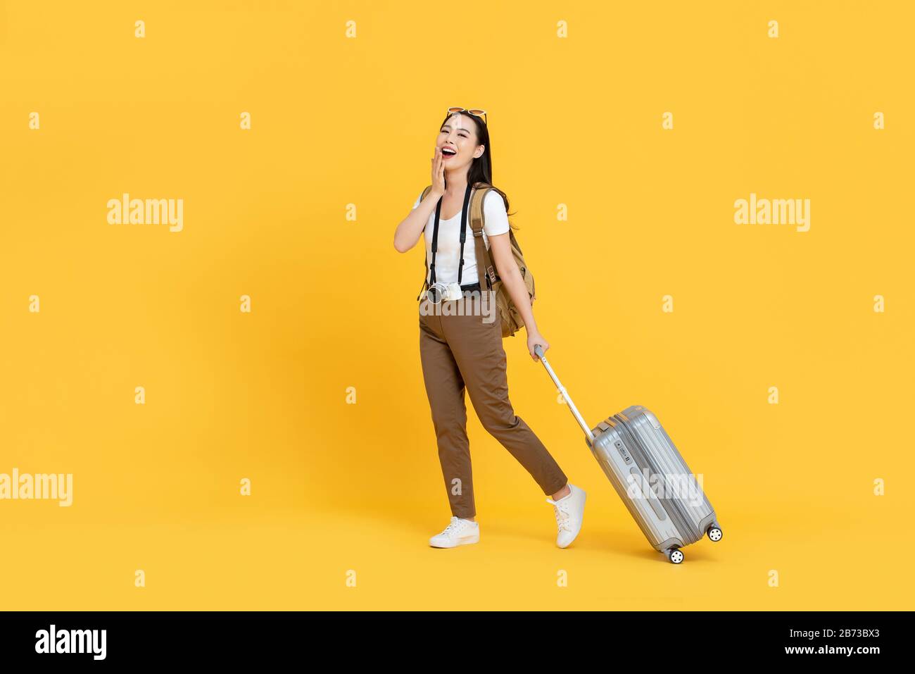 Fröhliche junge asiatische Touristin mit Gepäck, die an Feiertagen auf gelbem Hintergrund reisen wird Stockfoto