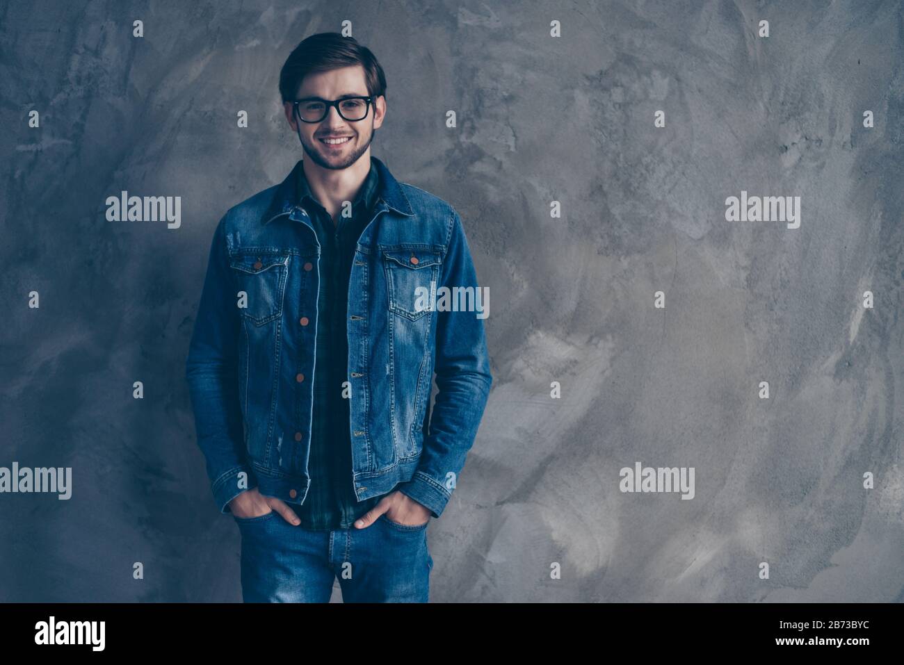 Fröhlicher, trendiger junger, gutaussehender Typ in Brille und mit einem Denimanzug, der auf grauem Hintergrund steht, lächelt und direkt auf die Kamera blickt. Stockfoto