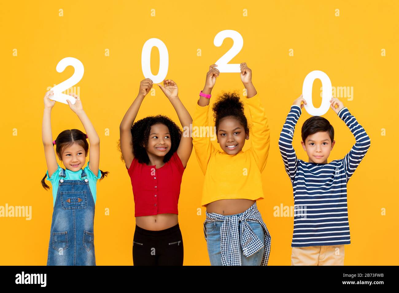 Niedliche gemischte Rennkinder lächeln und halten 2020-Zahlen für ein neues Jahreskonzept isoliert auf gelbem Hintergrund Stockfoto