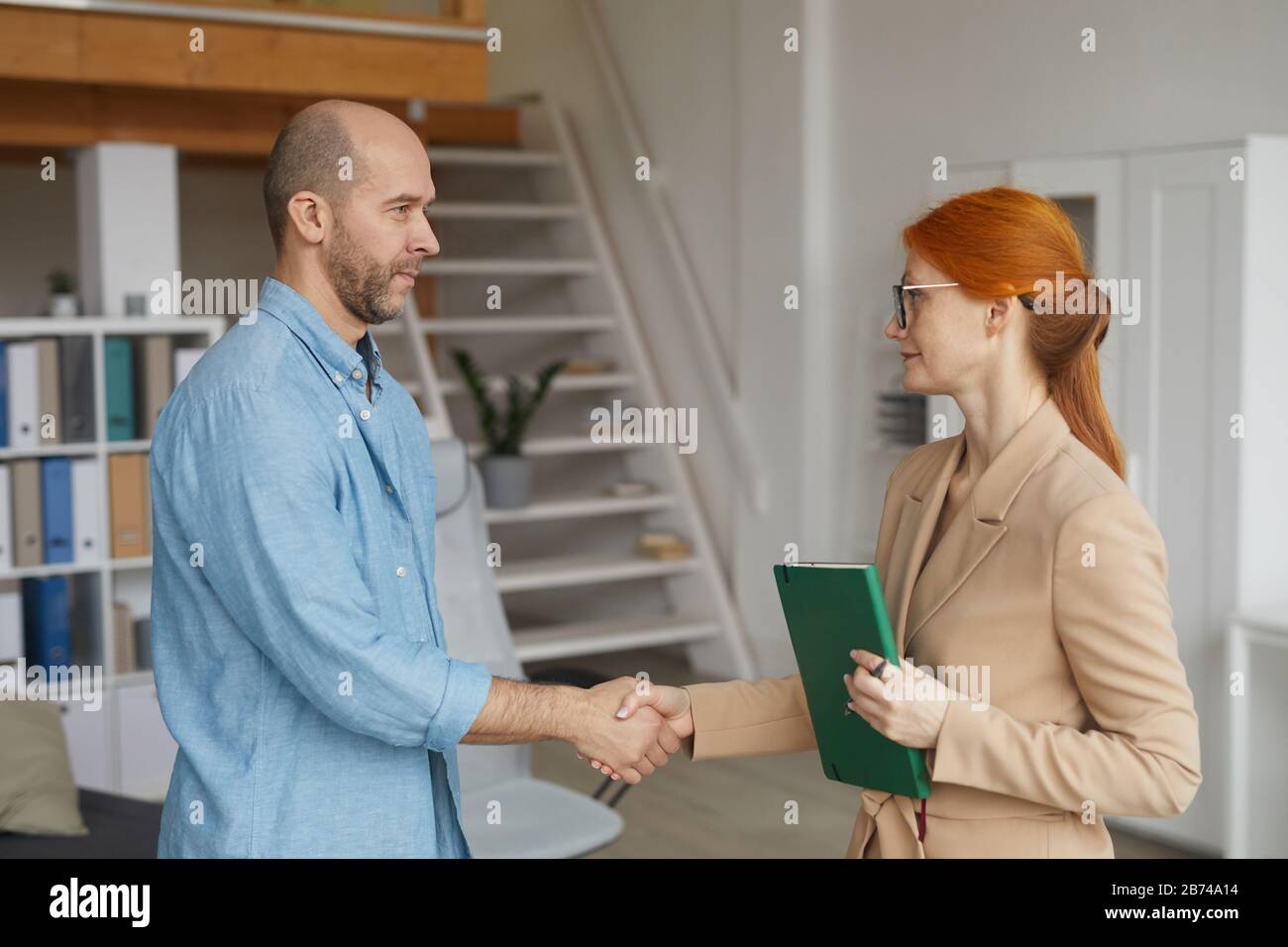 Junge rothaarige Geschäftsfrau in Brillen schüttelt mit Mann die Hände, während sie ein Treffen im Büro haben Stockfoto