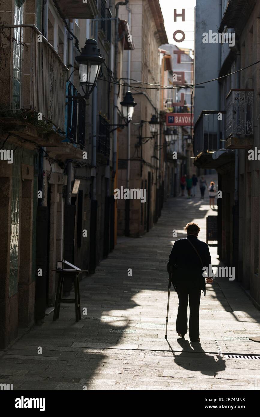 Eine ältere Frau, die einen Spazierstock benutzt, geht eine gepflasterte Gasse hinauf zu einem Hotel im alten Stadtgebiet von Baiona, Spanien Stockfoto
