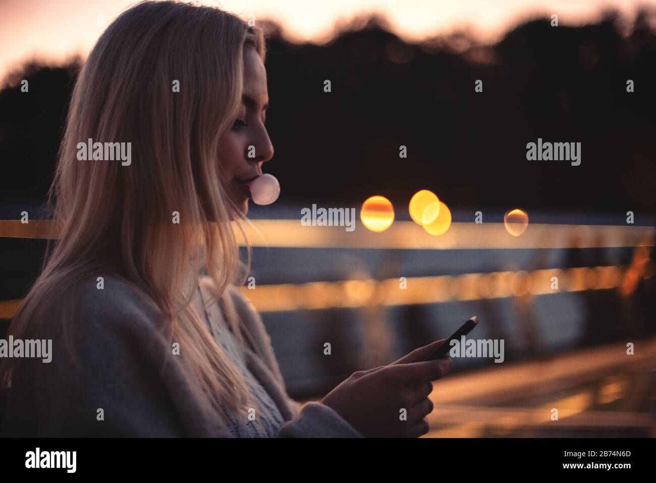 Junge Frau bläst während der Abenddämmerung mit dem Smartphone auf der Bank Stockfoto