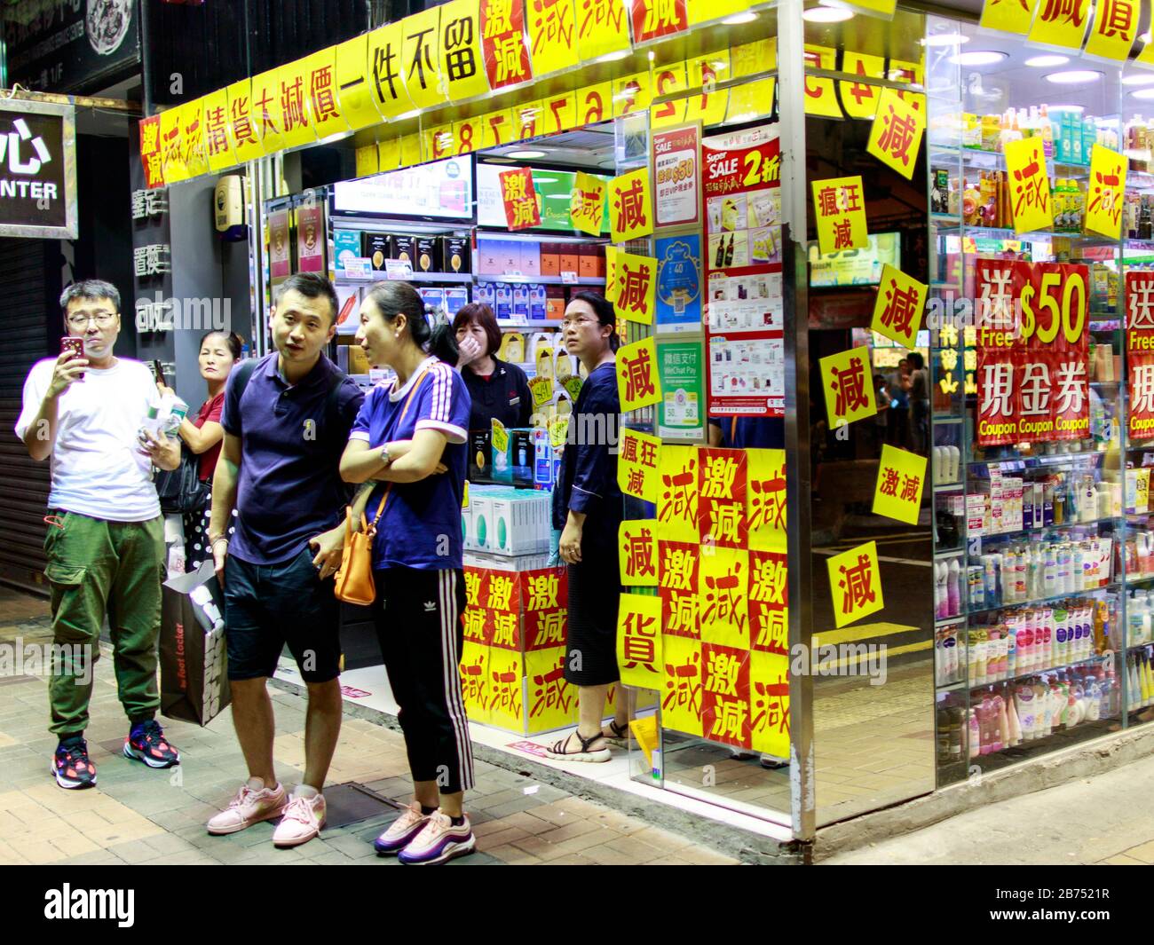 Touristen beobachten eine Kundgebung vor einem beliebten Laden von Chinesischen Festlandtouristen mit Rabattzeichen. Hongkongers protestieren auf der Straße, nachdem das Govenment heute ein Anti-Maske-Gesetz ausgibt. Stockfoto