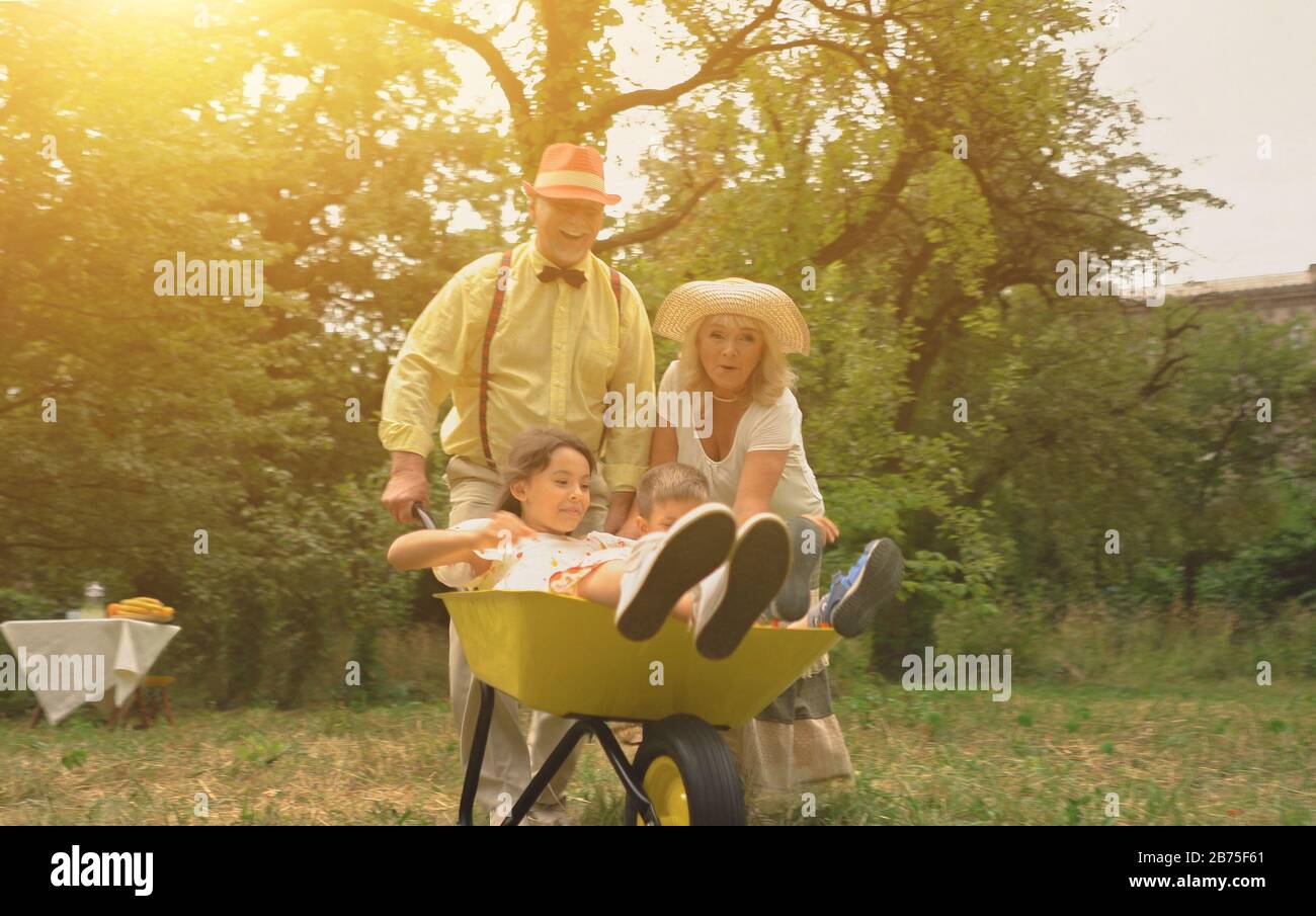 Das ältere Paar treibt seine Enkel in EINE Schubkarre.alle Familienmitglieder sind sehr glücklich.Konzept des aktiven Urlaubs Stockfoto