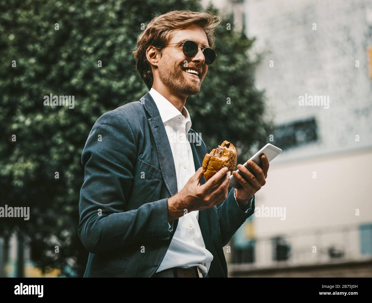 Junger weißer Kolar oder Freiberufler, der unterwegs ein Mittagessen hat. Gutaussehender Mann in einem blauen Sute und Sonnenbrille lächelnd mit Smartphone und Hamburger in seinem h Stockfoto