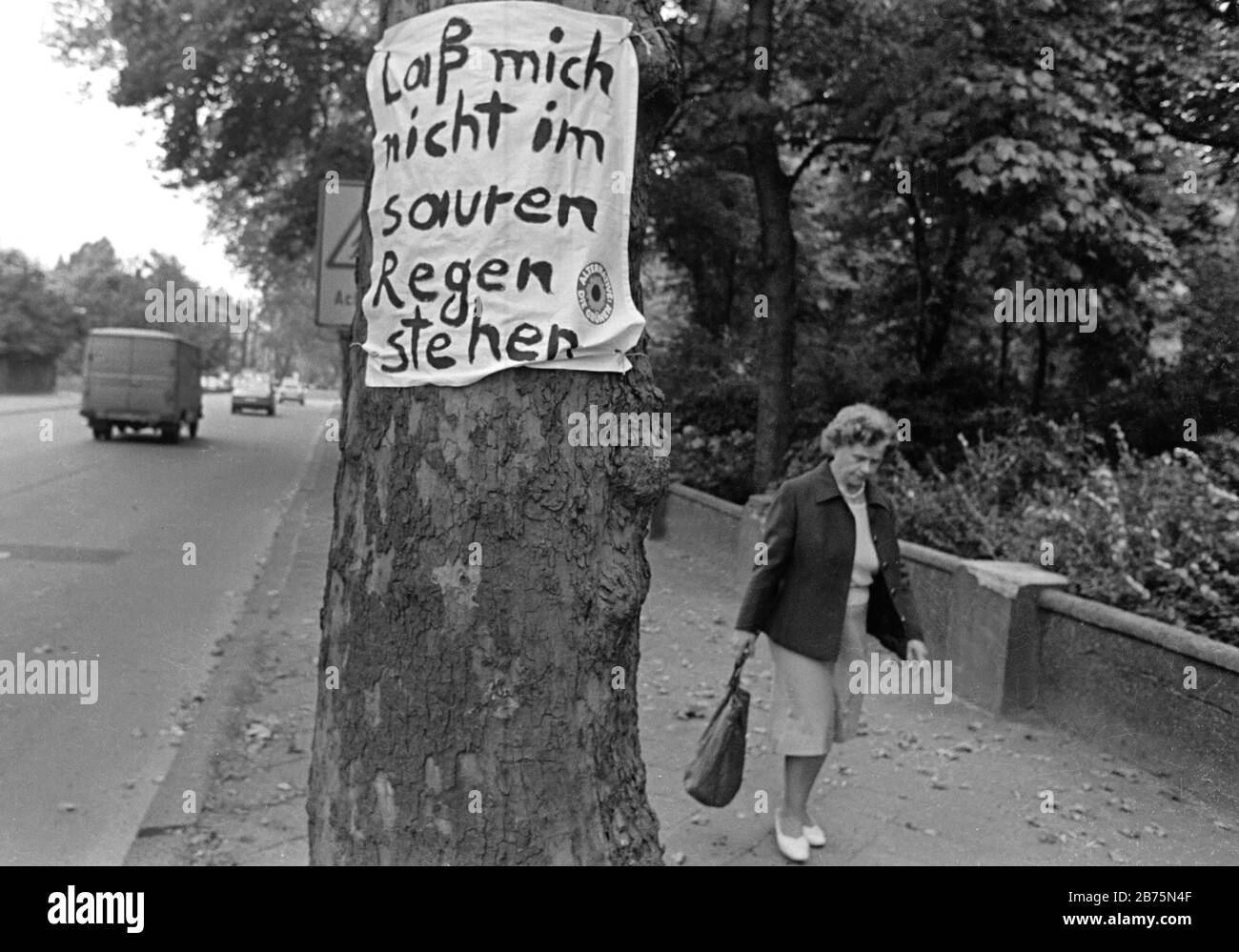 Eine Frau läuft an einem Baum auf einer Straße in Gelsenkirchen vorbei, wo ein Plakat der Partei "die Grünen" mit dem Text "Ich lasse mich nicht in saurem Regen stehen" angebracht ist, 19. Mai 1984. [Automatisierte Übersetzung] Stockfoto