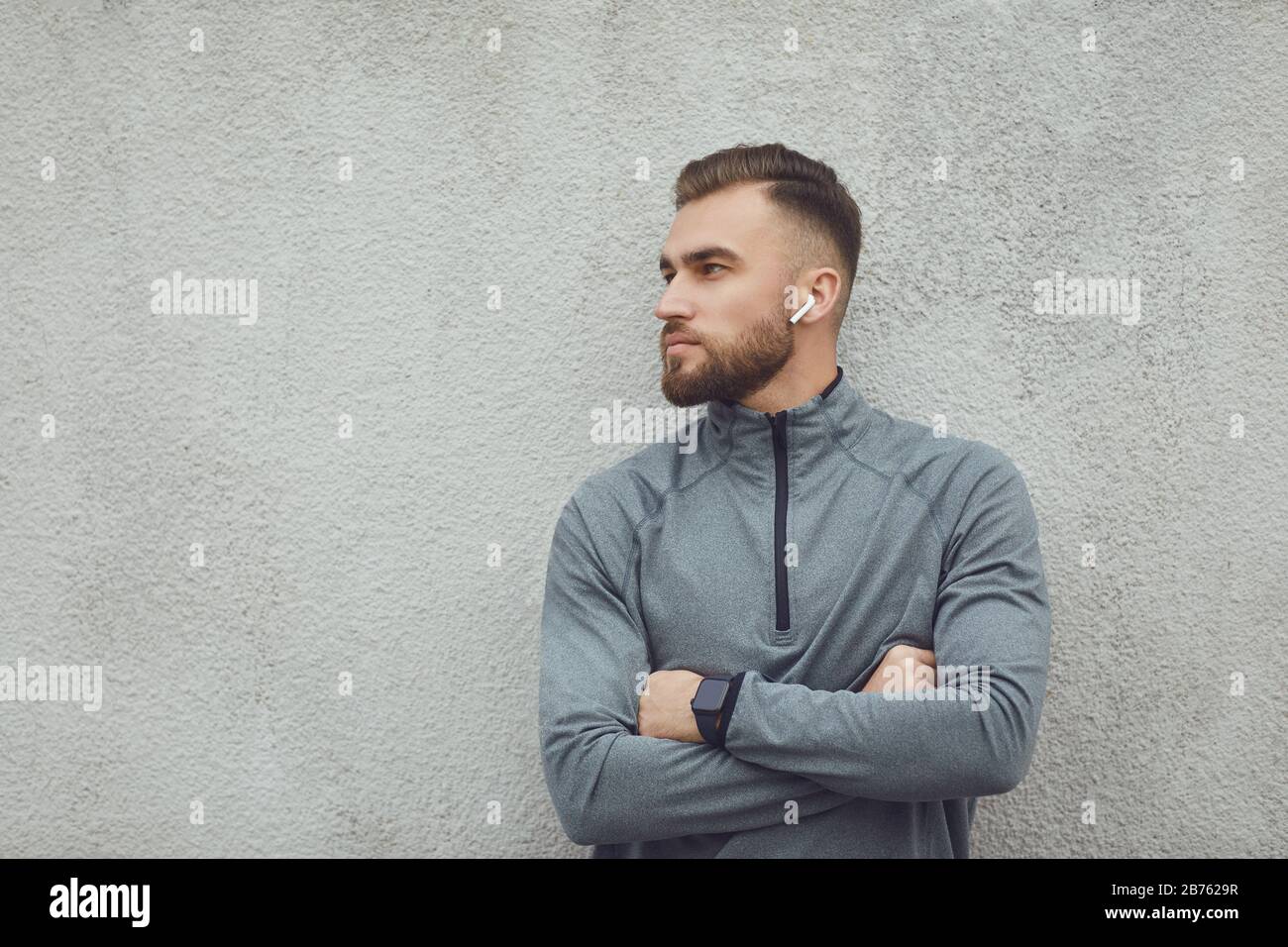 Bärtiger Kerl in grauer Sportswear mit Kopfhörern auf grauem Hintergrund. Stockfoto