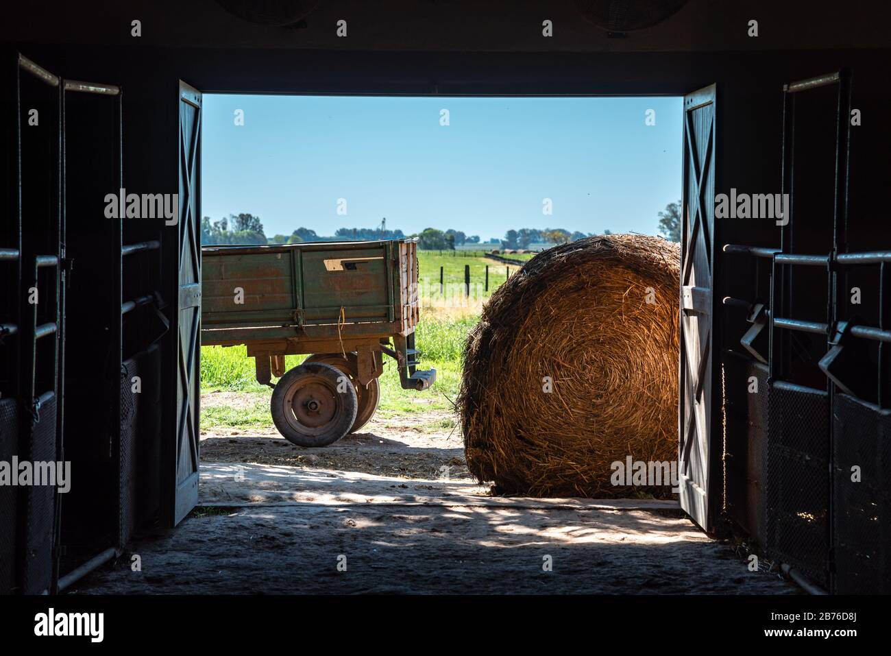 In einer Scheune, die nach draußen blickt, mit Heurolle oder Ballen und einem LKW-Anhänger mit blauem Himmel und grünem Gras Stockfoto In einer Scheune, die nach draußen blickt, mit Heurolle oder Ballen und einem LKW-Anhänger mit blauem Himmel und grünem Gras Stockfoto